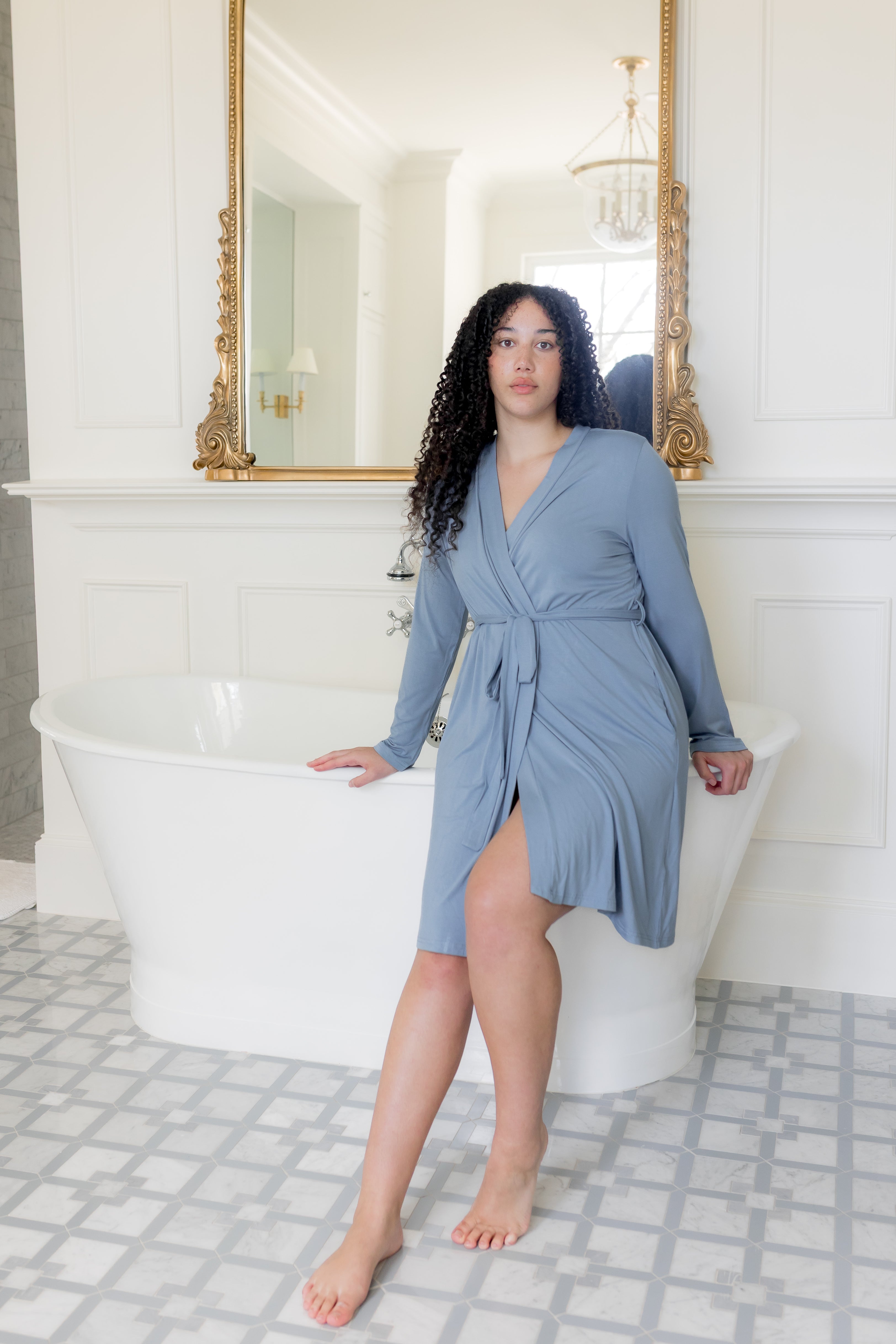 Woman in a blue dress standing in a bathroom with a white bathtub and tiled floor.