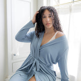Woman wearing a light blue robe in a bright room with large windows.