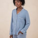Woman modeling slate blue button up pajamas with white piping against a tan backdrop with hand in pocket