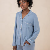 Woman modeling slate blue button up pajamas with white piping against a tan backdrop with hand in pocket