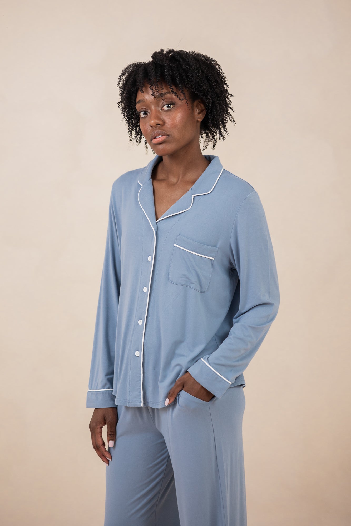 Woman modeling slate blue button up pajamas with white piping against a tan backdrop with hand in pocket