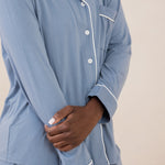 Woman modeling slate blue button up pajamas with white piping against a tan backdrop