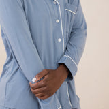 Woman modeling slate blue button up pajamas with white piping against a tan backdrop