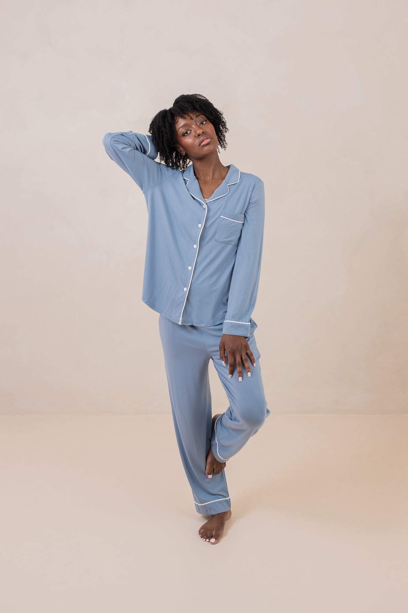 Woman modeling slate blue button up pajamas with white piping against a tan backdrop