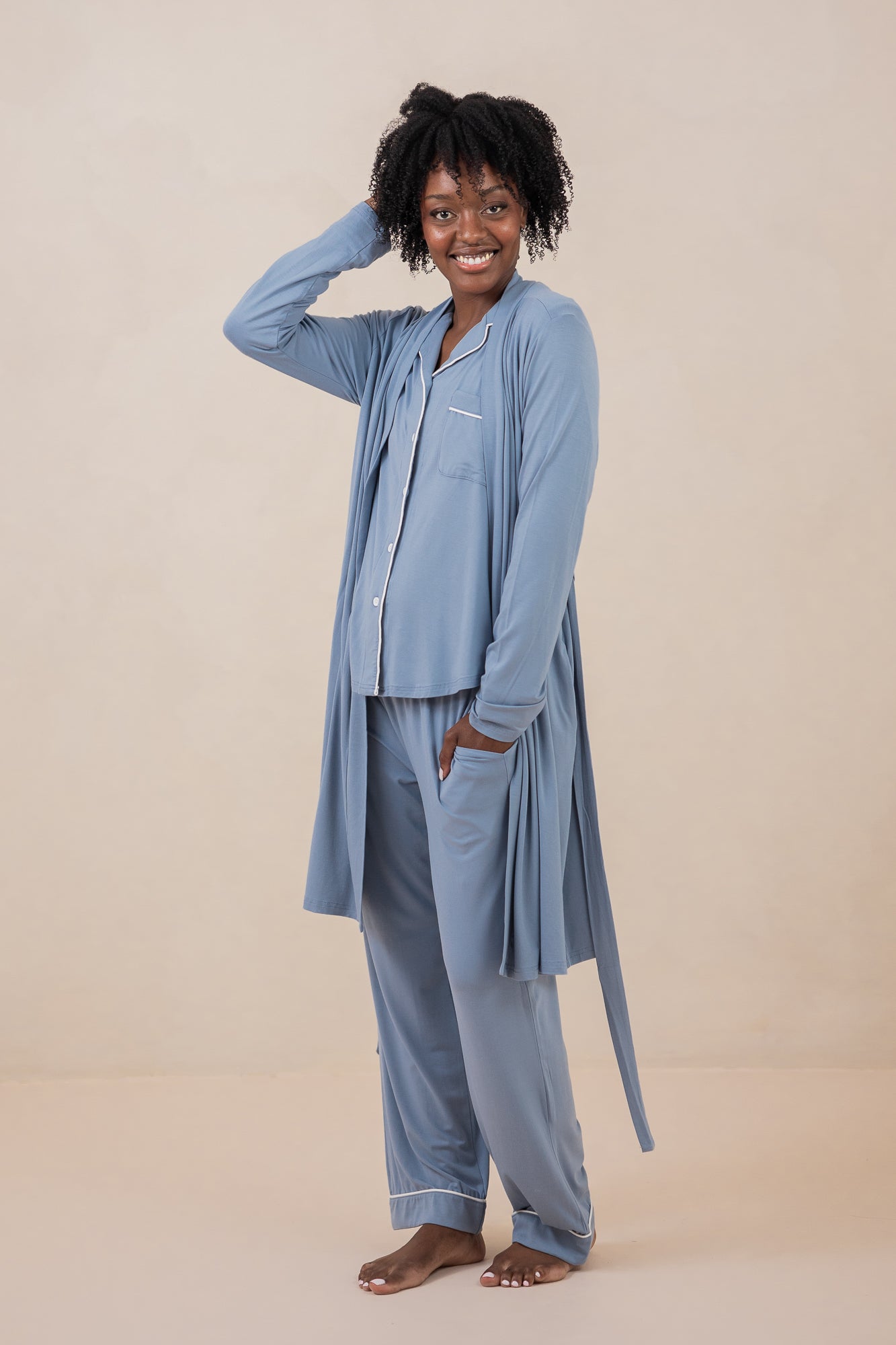 Woman modeling slate blue button up pajamas with white piping against a tan backdrop with matching slate robe