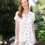 A woman wearing a short-sleeve button-front pajama top and matching shorts with a small floral print, standing outdoors on a garden path.