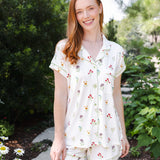 A woman wearing a short-sleeve button-front pajama top and matching shorts with a small floral print, standing outdoors on a garden path.