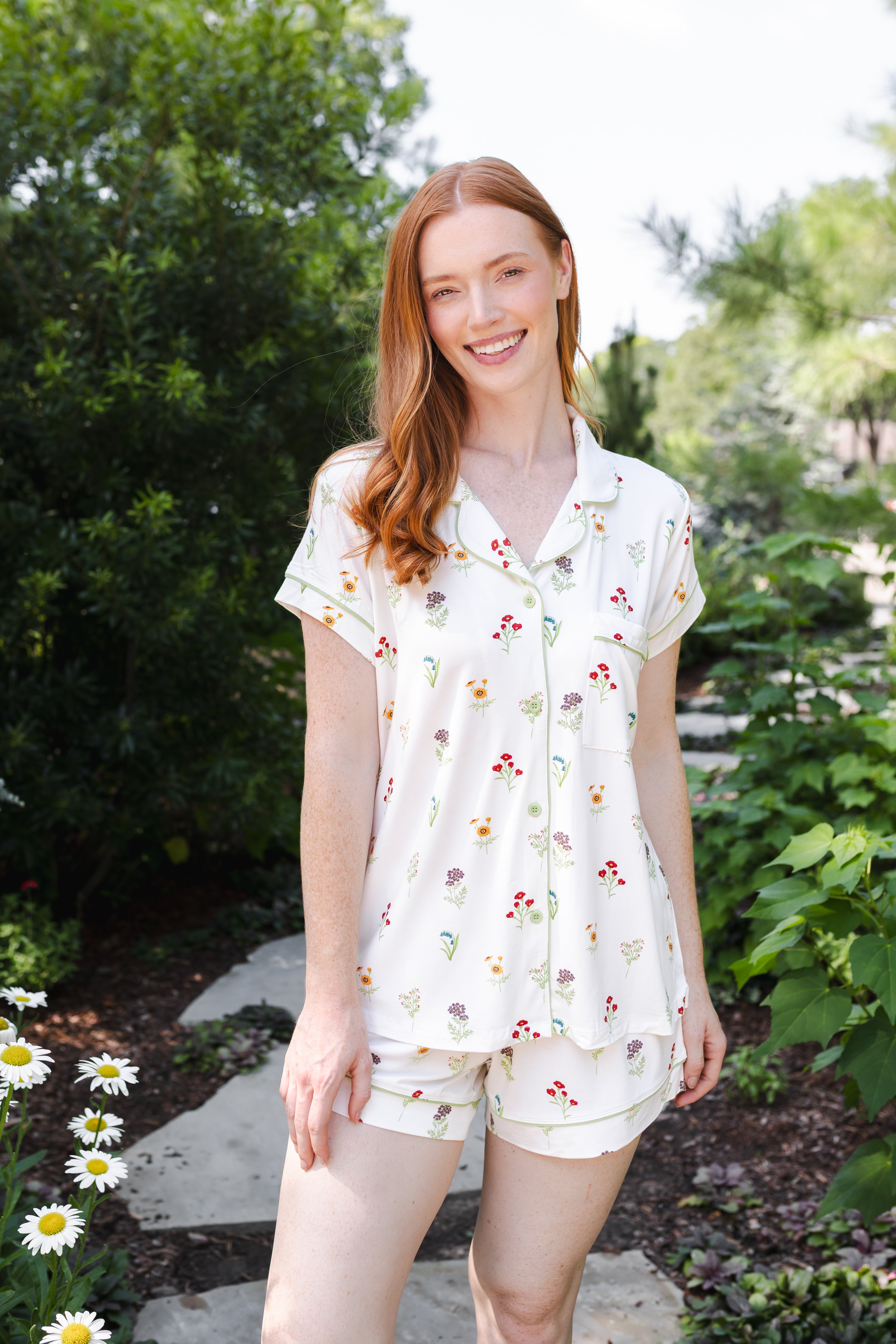 A woman wearing a short-sleeve button-front pajama top and matching shorts with a small floral print, standing outdoors on a garden path.