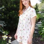 A woman with red hair wearing a short-sleeve button-front pajama shirt and matching shorts with a small floral print, standing in a garden path.