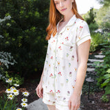 A woman with red hair wearing a short-sleeve button-front pajama shirt and matching shorts with a small floral print, standing in a garden path.