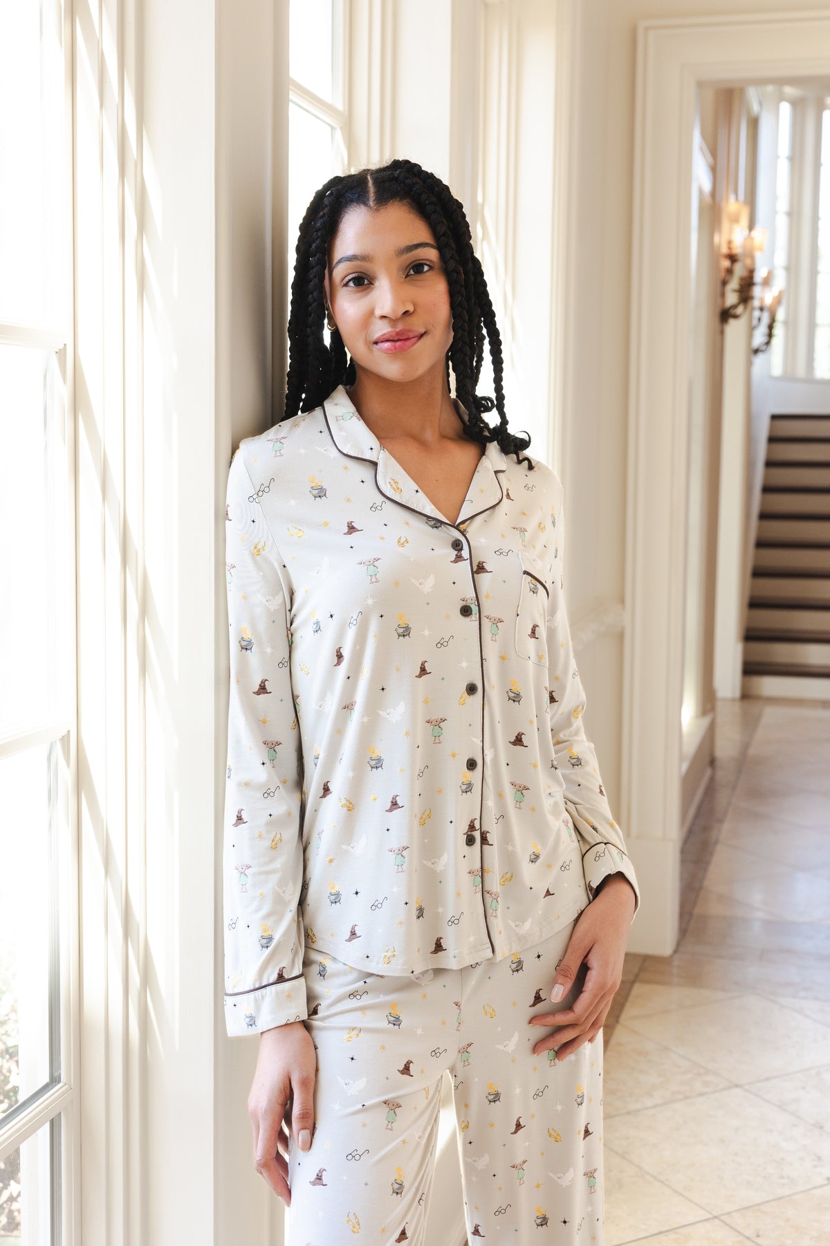 A woman wearing a matching pajama set with a whimsical Harry Potter pattern of small illustrated figures, standing in a brightly lit hallway with white walls and a staircase in the background.