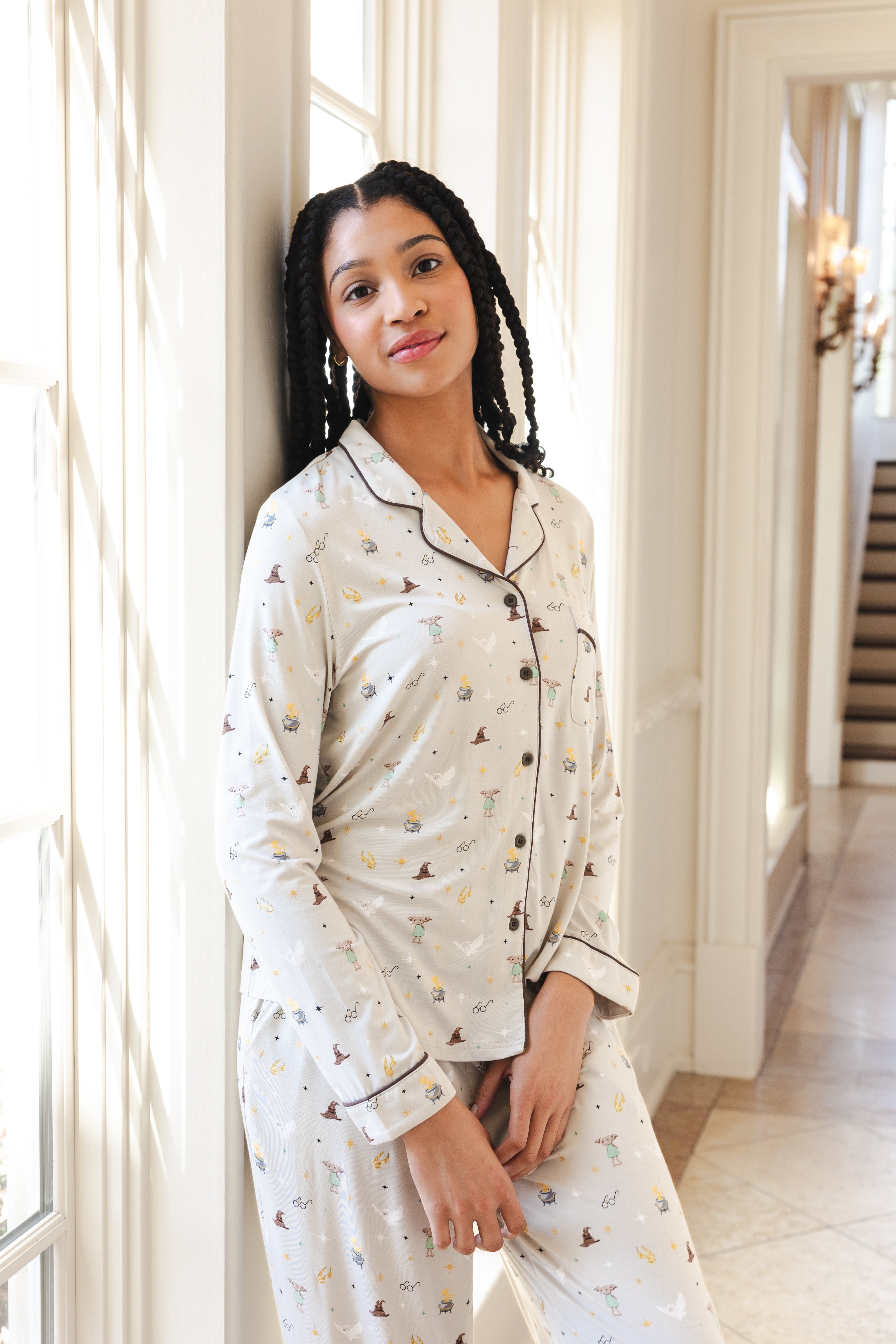 A woman wearing a matching long-sleeve pajama set with a small Harry Potter  pattern on a cream background, standing in a brightly lit hallway.