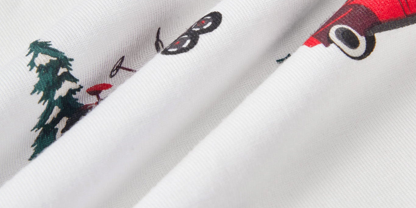 White fabric with a repeating print of red wagons, green pine trees, and a red sled, shown in a close-up folded view.