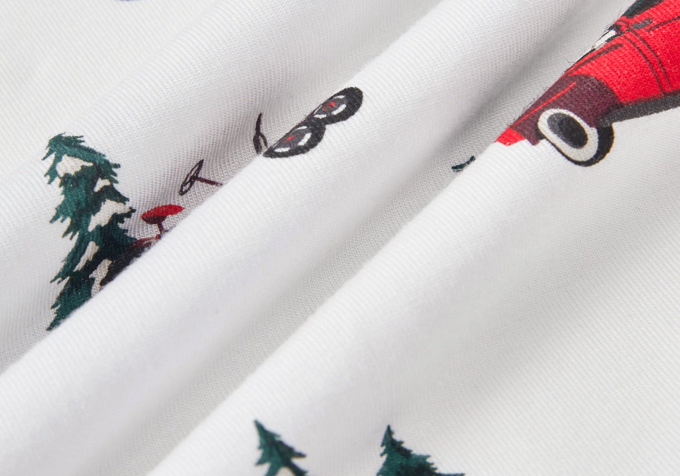 White fabric with a repeating print of red wagons, green pine trees, and a red sled, shown in a close-up folded view.
