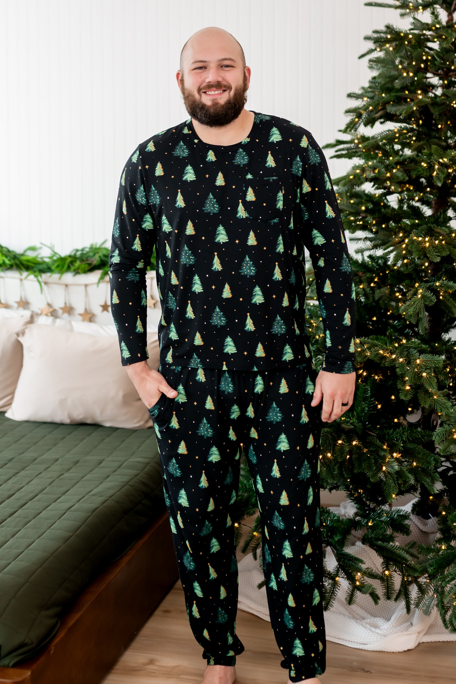 Smiling male model wearing the Men's Jogger Set in Twinkle Trees next to a Christmas tree and bed
