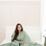 Model sitting on a neutral bed with Bamboo Blanket in Thyme 2.5 around her
