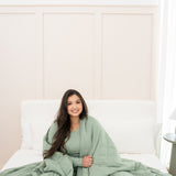 Model sitting on a neutral bed with Bamboo Blanket in Thyme 2.5 around her