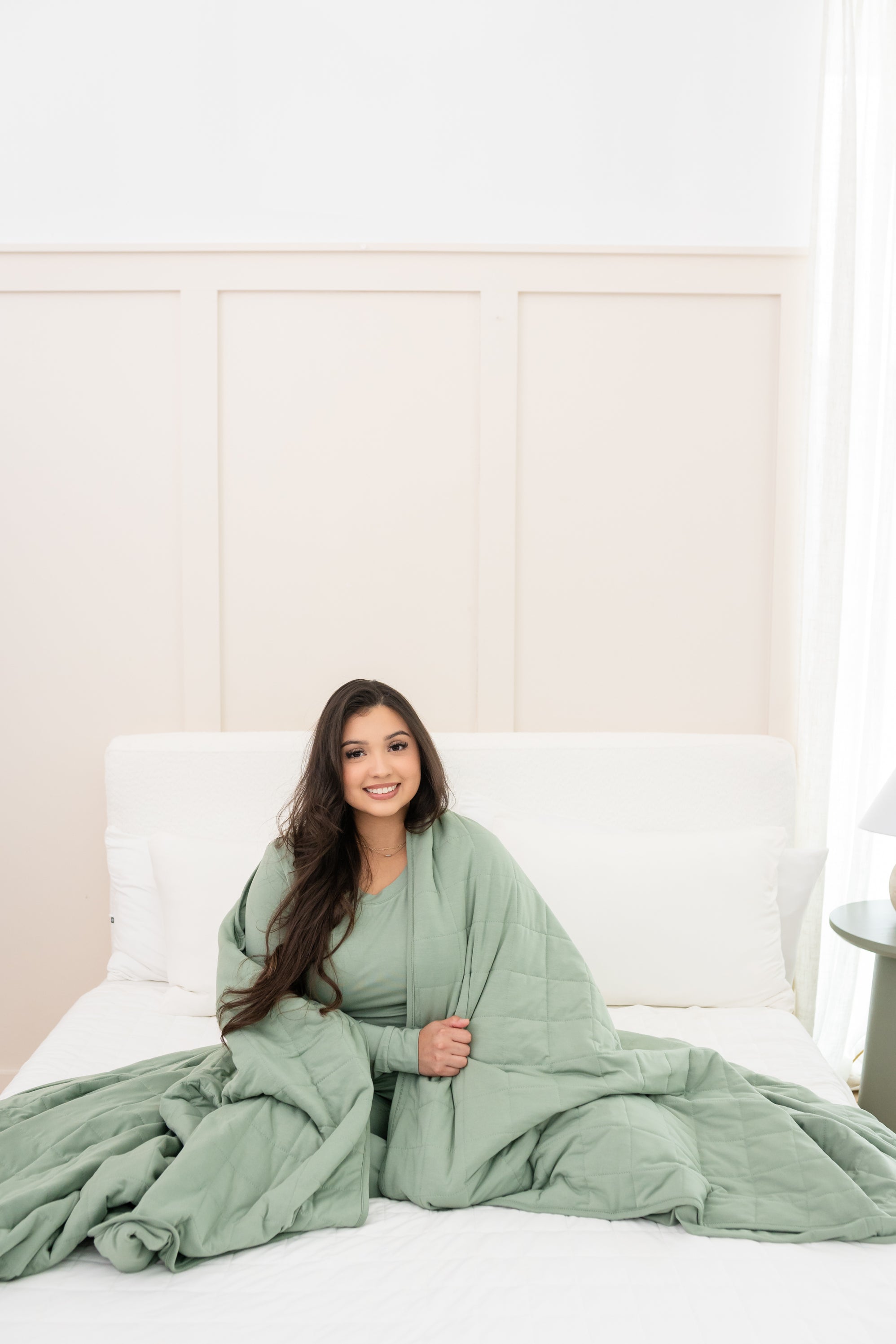 Model sitting on a neutral bed with Bamboo Blanket in Thyme 2.5 around her