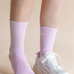 A close-up of a person's lower legs wearing light purple ribbed crew socks and a white chunky sneaker against a solid beige background.