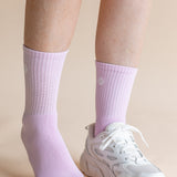 A close-up of a person's lower legs wearing light purple ribbed crew socks and a white chunky sneaker against a solid beige background.