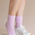 A close-up of a person's feet wearing light purple ribbed crew socks and a white athletic sneaker against a solid beige background.