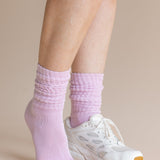 A close-up of a person's feet wearing light purple ribbed crew socks and a white athletic sneaker against a solid beige background.