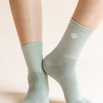 A close-up of a person's feet wearing ribbed crew socks in a pale sage green color, with a small white geometric logo on the side of the right sock.