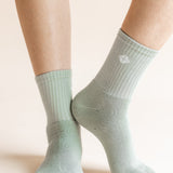 A close-up of a person's feet wearing ribbed crew socks in a pale sage green color, with a small white geometric logo on the side of the right sock.