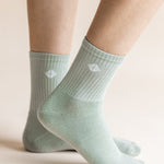 A close-up of a person's feet wearing light sage green ribbed crew socks with a small white geometric logo on the side, set against a warm beige background.