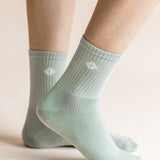 A close-up of a person's feet wearing light sage green ribbed crew socks with a small white geometric logo on the side, set against a warm beige background.