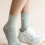 A close-up of a person's feet wearing pale sage green ribbed crew socks and white athletic sneakers against a soft beige background.