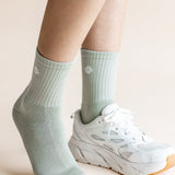 A close-up of a person's feet wearing pale sage green ribbed crew socks and white athletic sneakers against a soft beige background.