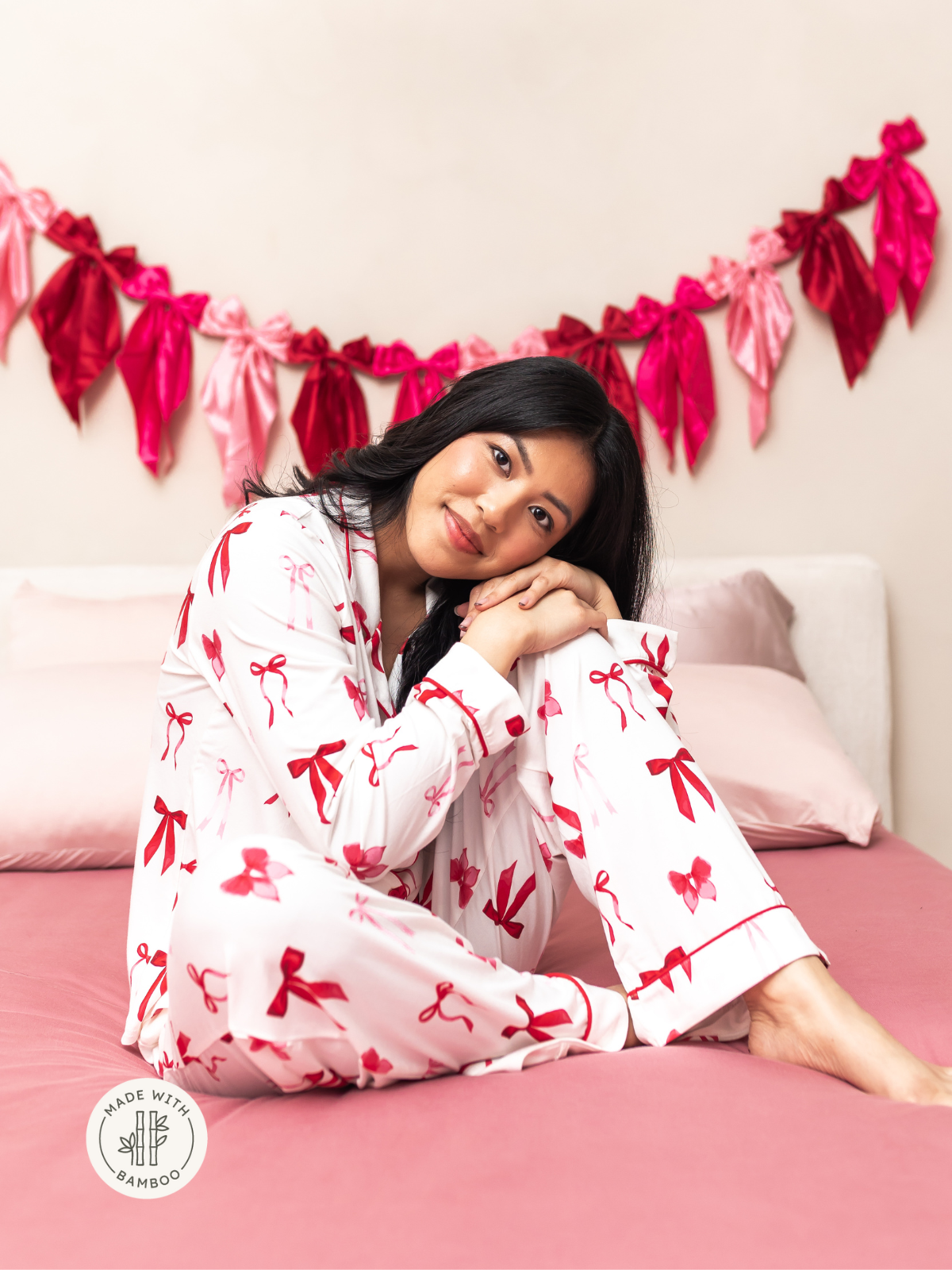Woman modeling white long sleeve pajamas with a red bow pattern while sitting on a bed with pink bamboo bedding