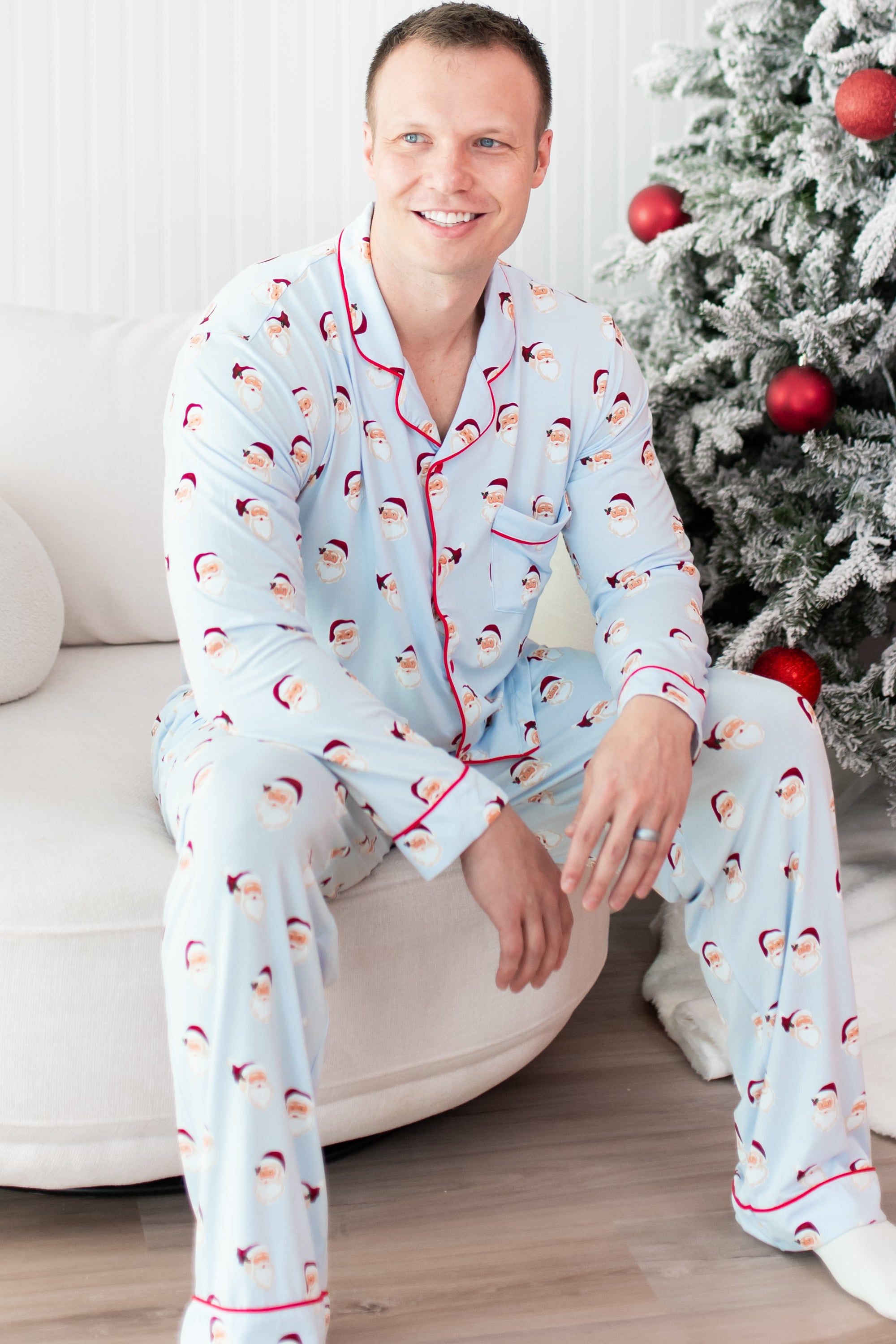 Man wearing light blue pajamas with santa pattern sitting on a white couch next to christmas tree