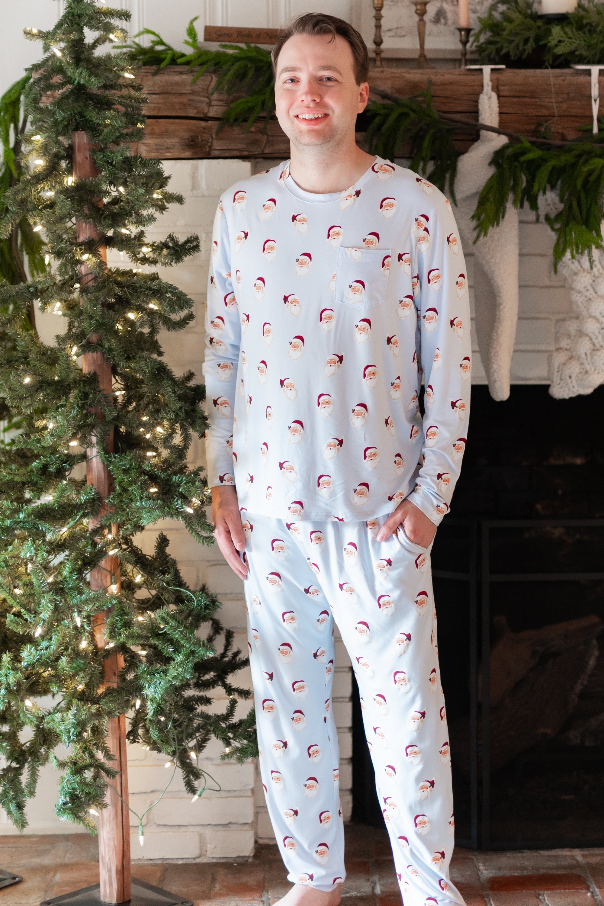 Smiling male model standing in front of a white brick fireplace with white stockings hung on the mantel wearing the Men's Jogger Set in Vintage Santa with one hand in the pants pocket
