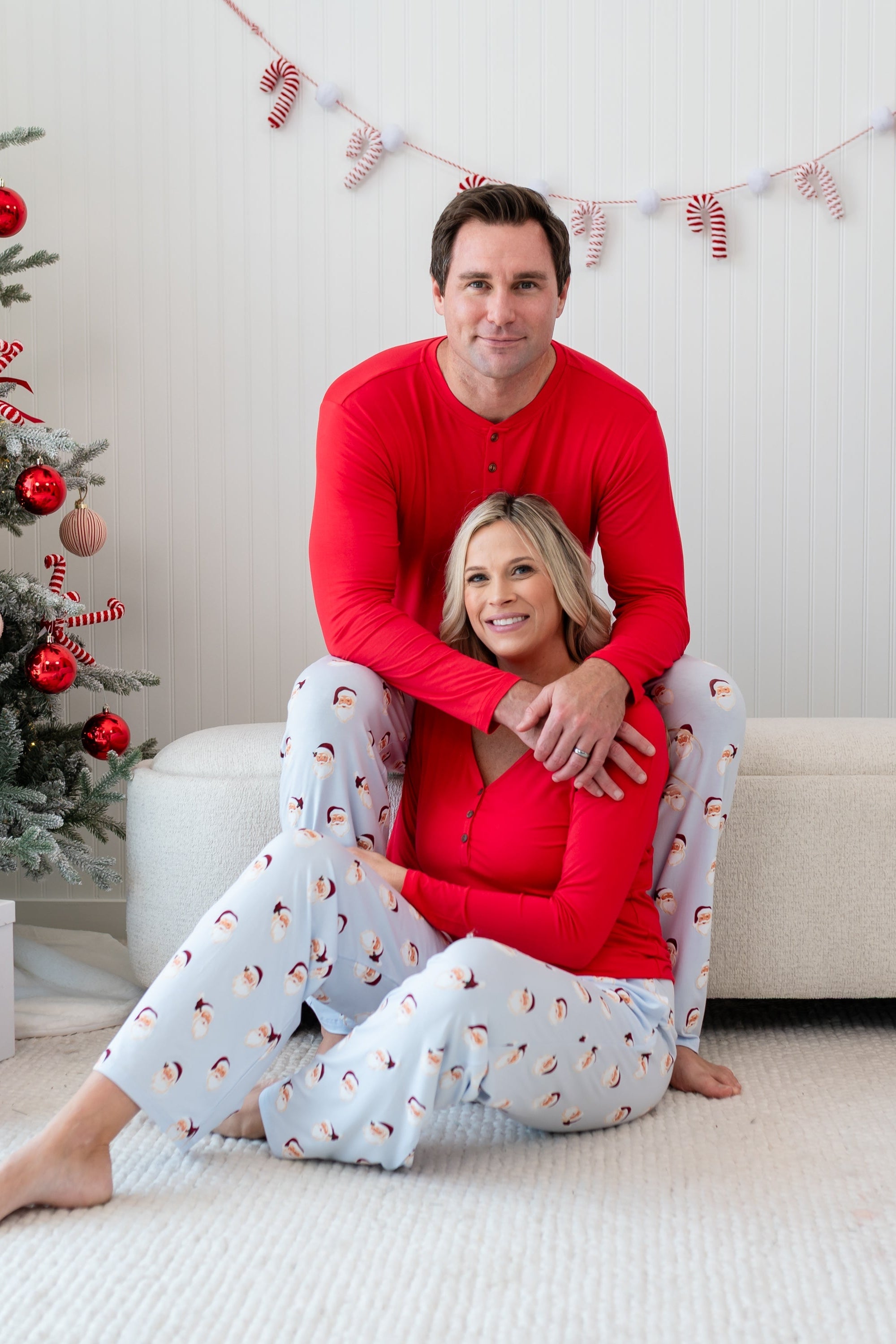 Husband wearing the Men's Lounge Pants in Vintage Santa and Cardinal Henley sitting on an ottoman with his wife in front of him matching in Vintage Santa Lounge pants and a Women's Cardinal Henley