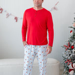 Male model standing in front of a cream colored ottoman and beside two decorated Christmas trees wearing the Men's Lounge Pants in Vintage Santa and a Men's Henley top in Cardinal