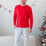 Male model standing in front of a cream colored ottoman and beside two decorated Christmas trees wearing the Men's Lounge Pants in Vintage Santa and a Men's Henley top in Cardinal