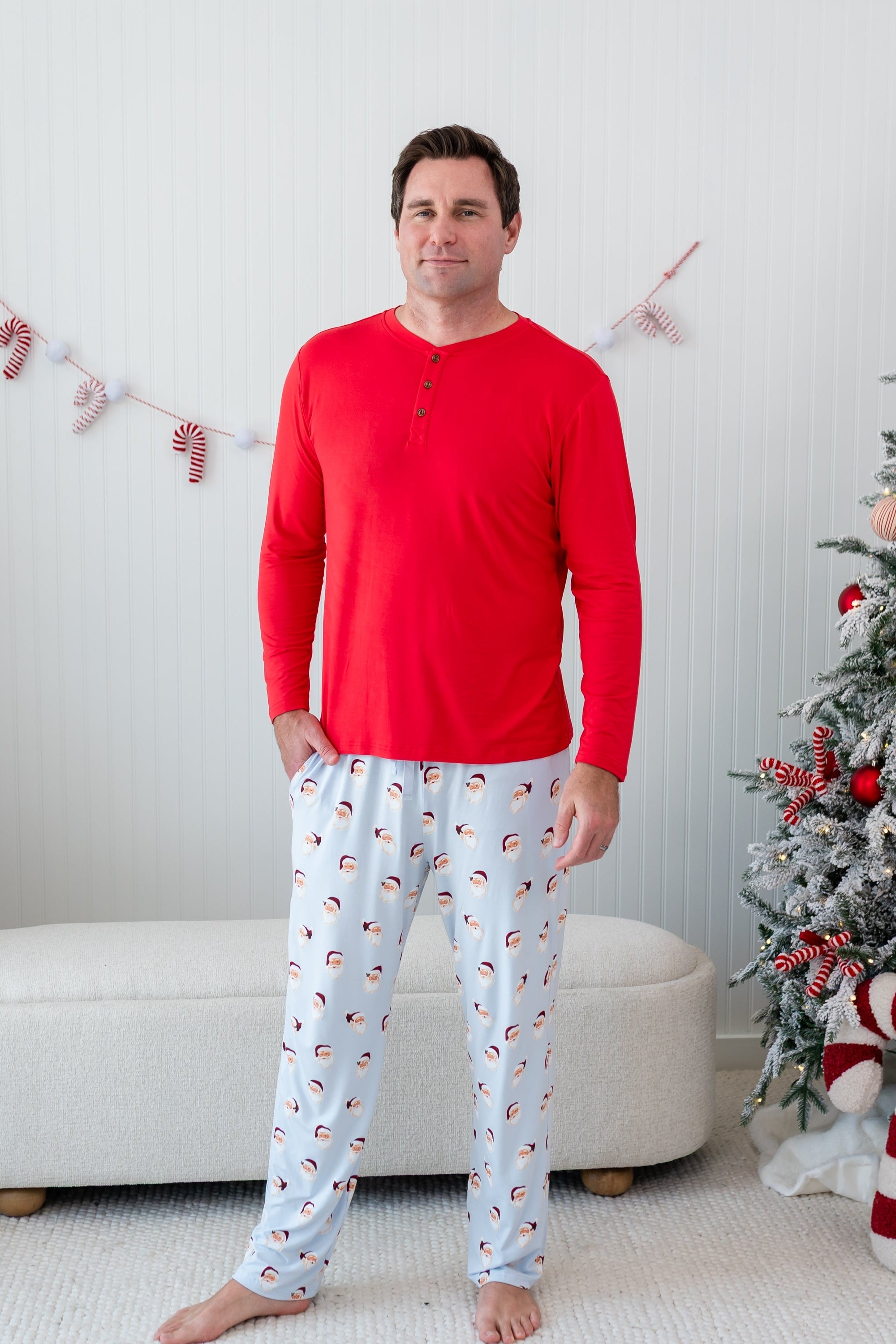 Male model standing in front of a cream colored ottoman and beside two decorated Christmas trees wearing the Men's Lounge Pants in Vintage Santa and a Men's Henley top in Cardinal