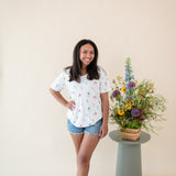Woman modeling Women’s Relaxed Fit V-Neck in Wildflower and denim shorts next to a floral arrangement  