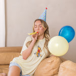 Woman sitting on a couch wearing a party hat and blowing into a noisemaker with balloons nearby.
