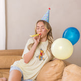Woman sitting on a couch wearing a party hat and blowing into a noisemaker with balloons nearby.