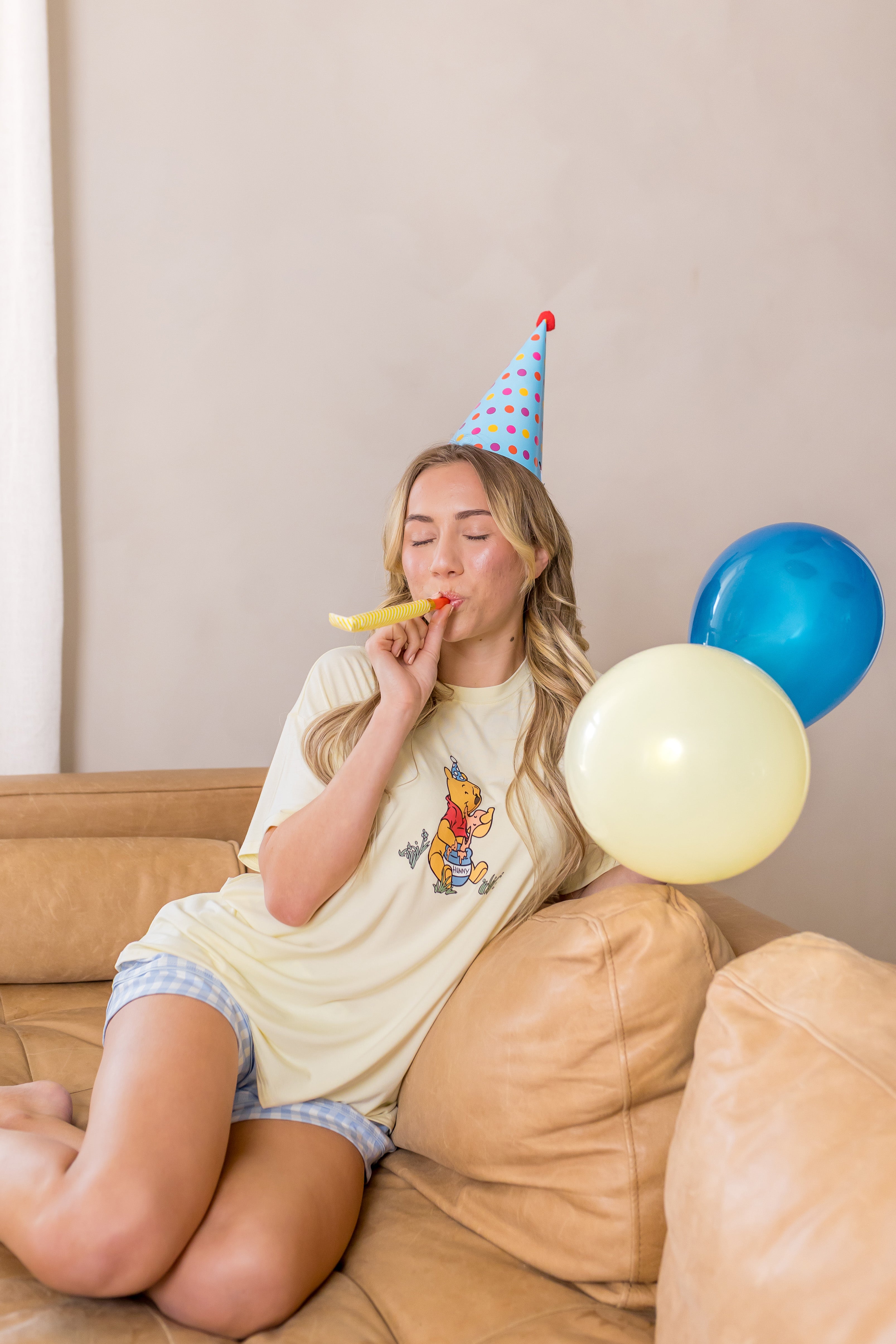 Woman sitting on a couch wearing a party hat and blowing into a noisemaker with balloons nearby.