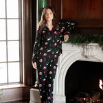 A woman wearing a black long-sleeve pajama set with a Santa Claus and snowman print, standing barefoot in front of a fireplace with a window to her left.
---1605 3