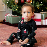 A toddler sitting on a patterned red rug in front of a decorated Christmas tree, wearing a black long-sleeve pajama set with a Santa Claus and snowman print.