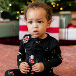 A toddler sitting on a patterned rug in front of a decorated Christmas tree and wrapped gifts, wearing a black long-sleeve pajama set with a festive holiday print.