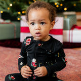 A toddler sitting on a patterned rug in front of a decorated Christmas tree and wrapped gifts, wearing a black long-sleeve pajama set with a festive holiday print.