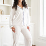 Woman wearing white pajamas with black trim in a room with white walls and wooden floor.