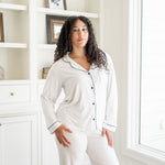 Woman wearing a white pajama set in a room with a bookshelf and window.