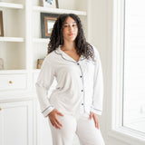 Woman wearing a white pajama set in a room with a bookshelf and window.